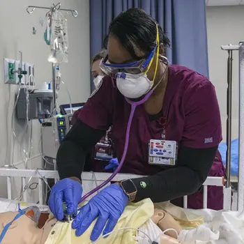 Nursing student in scrubs with stethoscope listening to simulated patient