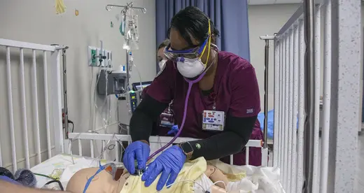 Nursing student in scrubs with stethoscope listening to simulated patient