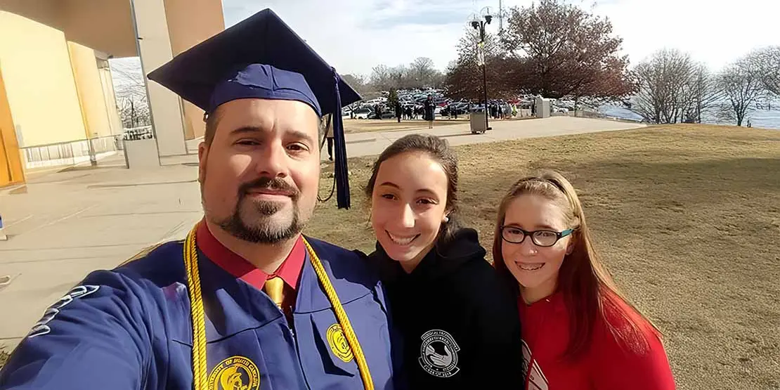 Daniel Stokoe wearing commencement gown with daughters Kayla in black hoodie and Kira in red.